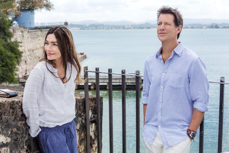 In a still from "Stranded in Paradise", Tess and Carter stand looking off camera in front of a Puerto Rico ocean backdrop. 