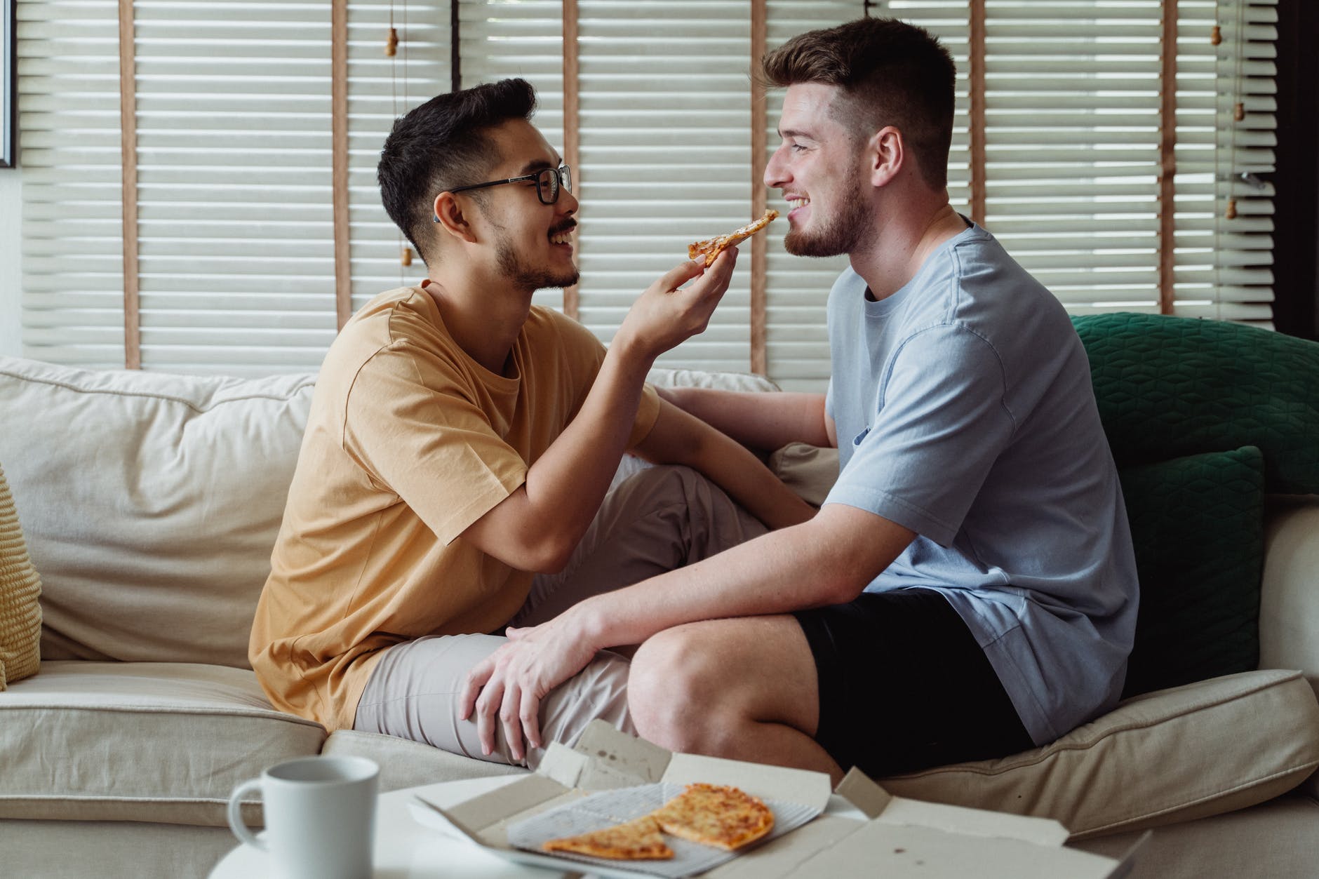 Male couple sit on couch, feeding each other pizza on Valentine's Day. 